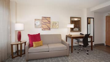 A hotel room with a sofa, a desk, a chair, a lamp, and artwork on the wall.