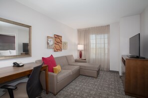 A hotel room with a desk, a sofa, a TV, a lamp, and a mirror.