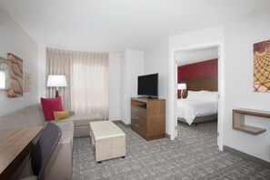 Quarto de hotel com cama, televisão, escrivaninha, cadeira, abajur, mesa de cabeceira e um pequeno armário.