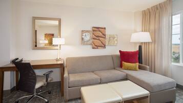 A hotel room with a desk, office chair, sofa, ottoman, and a large window with curtains.