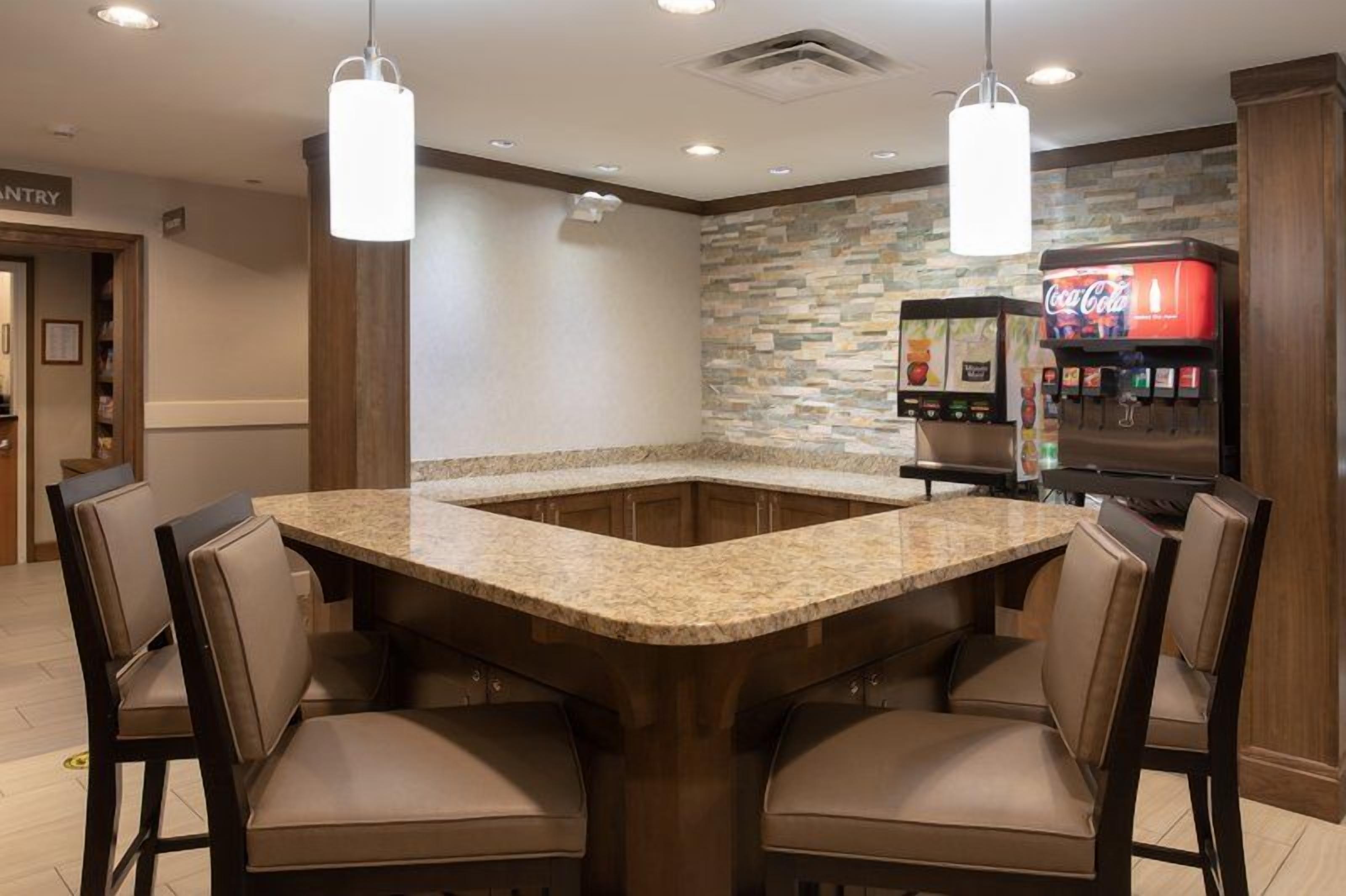 A kitchen area with a granite countertop, four chairs, a Coca-Cola vending machine, and a stone backsplash.