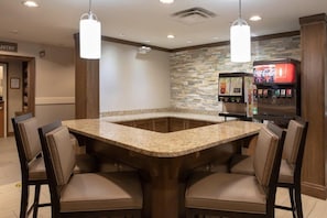A kitchen area with a granite countertop, four chairs, a Coca-Cola vending machine, and a stone backsplash.