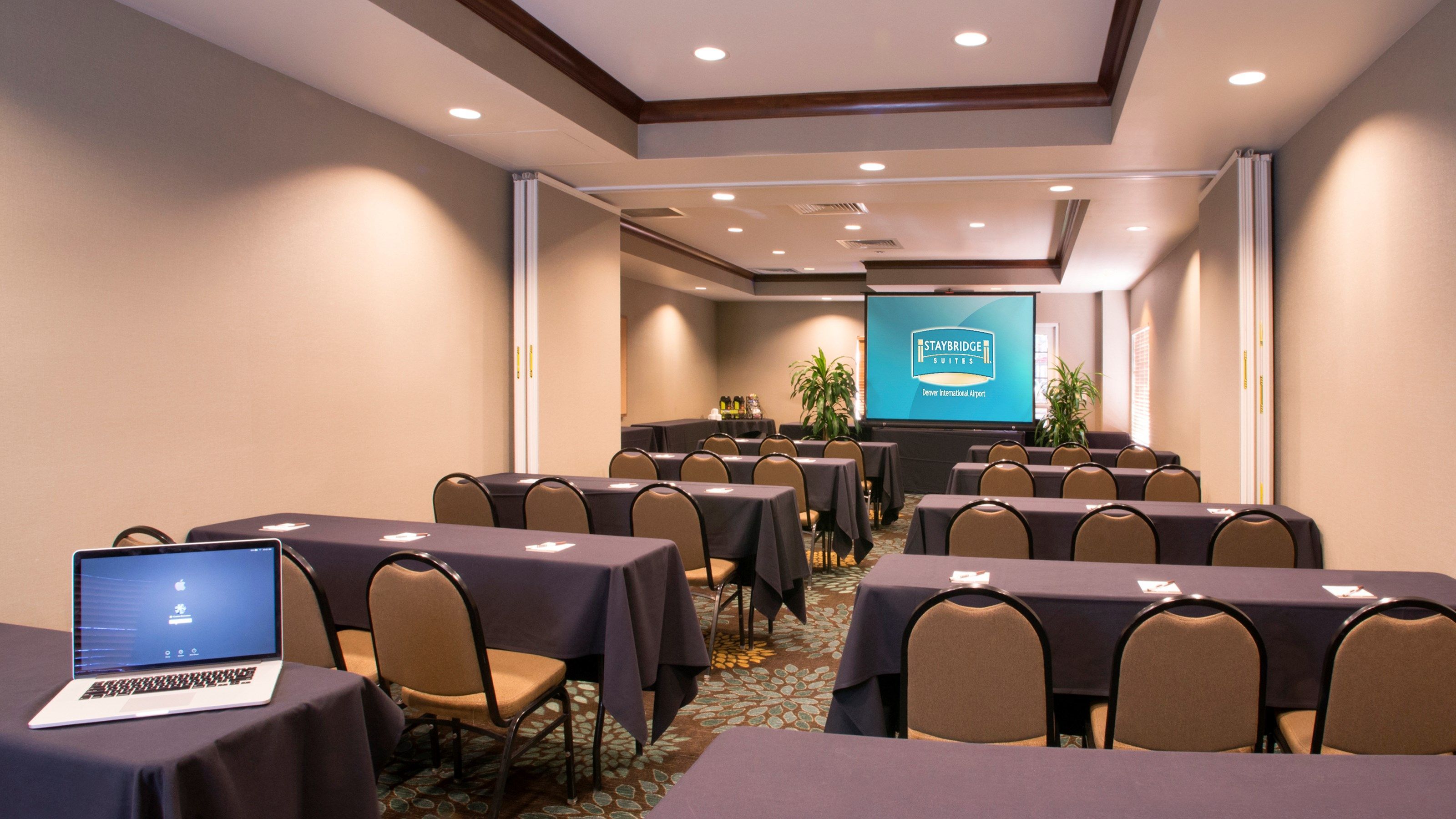 A conference room with tables, chairs, a laptop, and a presentation screen.