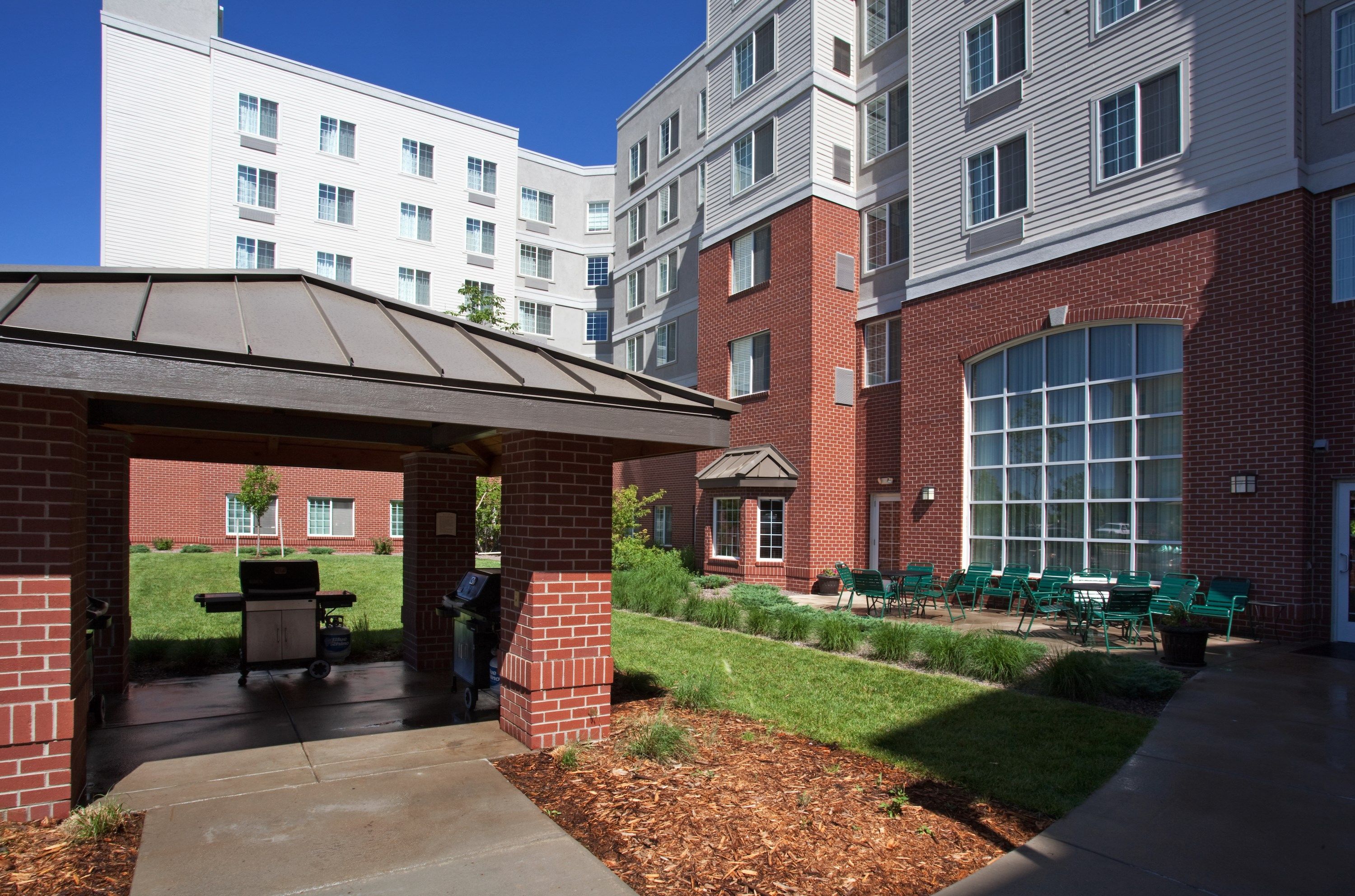 A covered outdoor area with a grill, brick building, and green chairs.