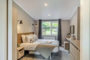 A bedroom with a bed, bedside tables, a desk, a radiator, a window with curtains, and a view of the outdoors. - Elwy 3.0 Spa (Denbigh)