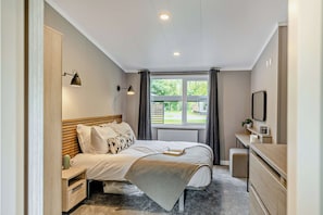 A bedroom with a bed, bedside tables, a desk, a radiator, a window with curtains, and a view of the outdoors. - Elwy 3.0 Spa (Denbigh)