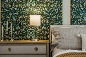 A bedroom with a floral wallpaper, a bedside lamp, a nightstand with a drawer, and a bed with a pillow. - The Landing | Bike to Downtown + Coler (Bentonville)