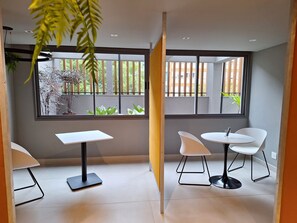 A modern room with a white table and chairs, large windows with a view of greenery, and a plant in the corner. - Modern studio apartment 200m from Paulista Avenue with a closed bedroom. (SÃO PAULO)