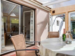 A room with a sliding glass door leading to an outdoor area, a round table with a tablecloth, wicker chairs, and a bottle of wine. - 1 in the House Sturmmowe (St. Peter-Ording)