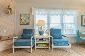 Une pièce avec deux fauteuils bleus, une petite table avec une lampe, un fauteuil en osier et une fenêtre avec des stores.