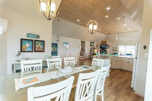 A spacious kitchen with wooden flooring, white cabinetry, and a dining area with white chairs and a glass table. - Vintage Mermaid Cottage. 350ft from Ocean. Hot Tub! (Corolla)