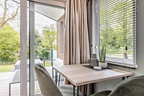 A modern outdoor seating area with a table and chairs, a view of greenery, and a sliding glass door. - Prion 1.0 Pet (Denbigh)
