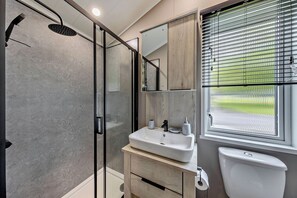 A modern bathroom with a glass-enclosed shower, a sink with a mirror above, and a toilet with a window above it. - Prion 1.0 Pet (Denbigh)