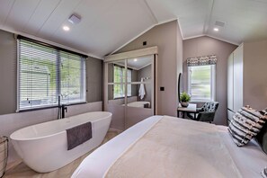 A modern bathroom with a freestanding bathtub, a large mirror, and a window with blinds. - Prion 1.0 Pet (Denbigh)