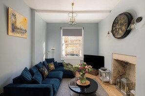 A living room with a blue sofa, a fireplace, a clock, and a painting on the wall. - 3-Bed Home in Batheaston, Sleeps 6 (Batheaston)