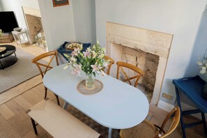A dining area with a white table, four wooden chairs, a vase with flowers, a blue side table, and a fireplace. - 3-Bed Home in Batheaston, Sleeps 6 (Batheaston)