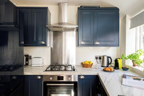 A modern kitchen with dark cabinets, a stainless steel range hood, a gas stove, an oven, a toaster, and a window with a view. - 3-Bed Home in Batheaston, Sleeps 6 (Batheaston)