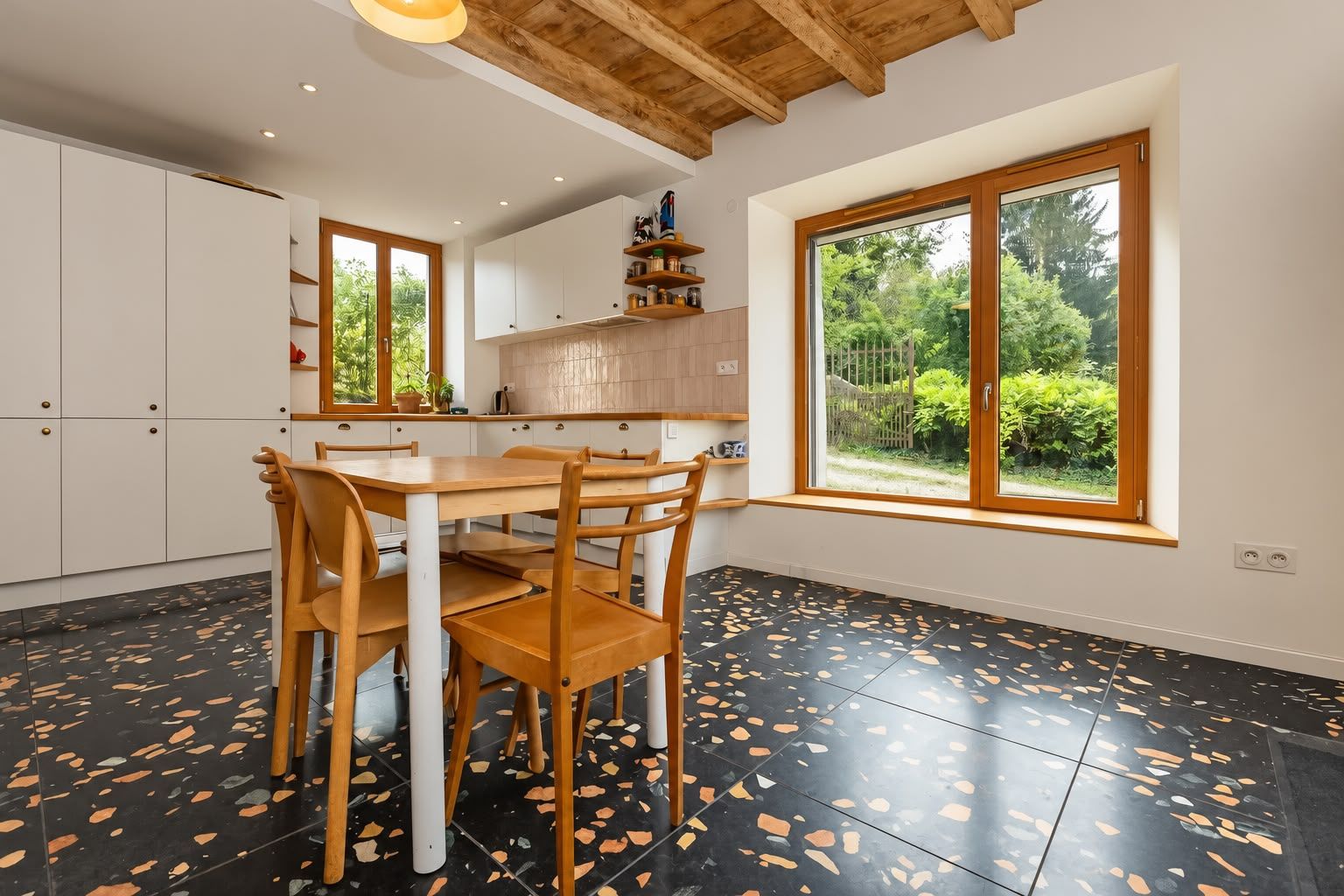Cocina moderna con armarios blancos, una mesa de comedor de madera y sillas. Se ve un ventanal con vistas a follaje exterior.