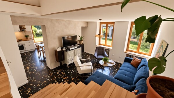 Un salon moderne avec un canapé bleu, une table basse blanche et un meuble pour la télévision. Un escalier mène à un autre niveau, et on aperçoit un coin cuisine au fond de la pièce.