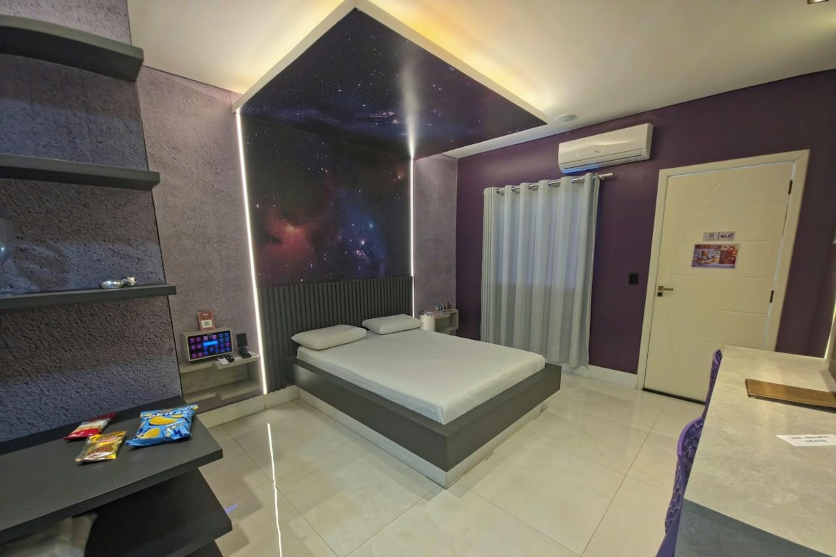 Un dormitorio con cama, mesita de noche, estante con aperitivos, aire acondicionado de pared y una puerta.