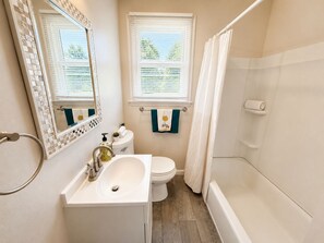 A bathroom with a white sink, toilet, and bathtub. There are two windows with blinds, and a towel rack with towels. - Southern Serenity - Cozy 2 BR • Near Nashville (La Vergne)