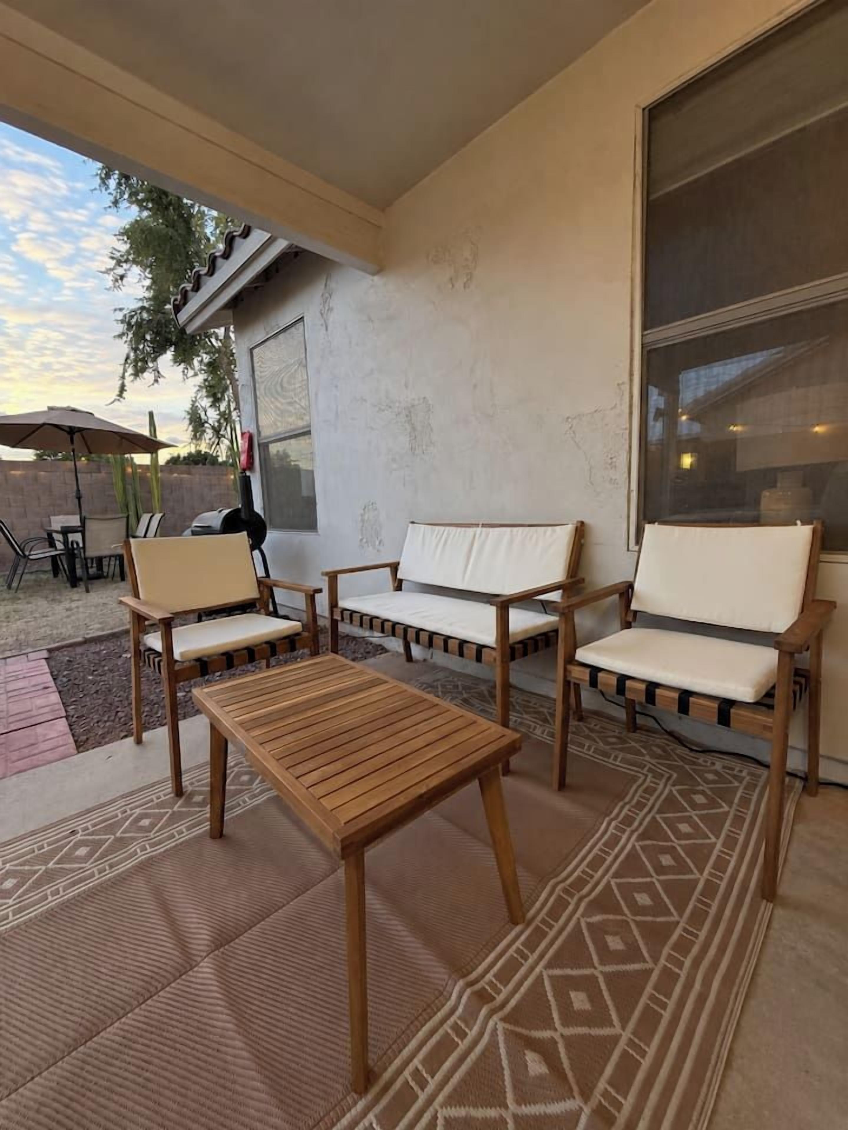 An outdoor patio with a wooden table and two cushioned chairs, a patterned rug, and a large window.