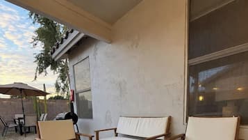 An outdoor patio with a wooden table and two cushioned chairs, a patterned rug, and a large window.