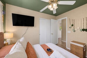 A hotel room with a bed, a television, a ceiling fan, a door, and a wall-mounted shelf with decorative items.