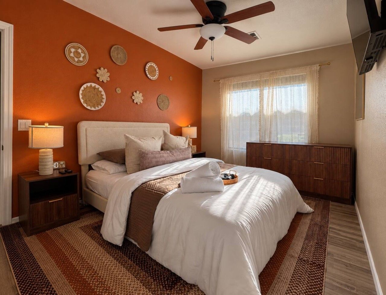 A bedroom with a bed, nightstands, a ceiling fan, a window with curtains, and wall decor.
