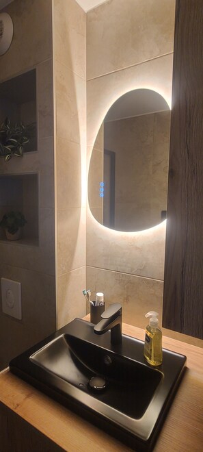 Ein modernes Badezimmer mit einem schwarzen Waschbecken, einem runden Spiegel mit indirekter Beleuchtung und einer kleinen Pflanze an der Wand.