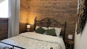 Een slaapkamer met een houten wand, een metalen bed, beddengoed met een patroon, twee kussens, een nachtkastje met een lamp en een raam met gordijnen.