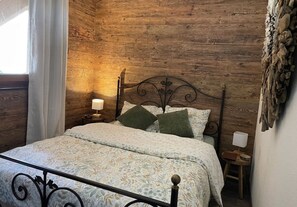Ein Schlafzimmer mit Holzwand, Metallbettgestell, einem Bett mit gemusterten Bettbezügen, zwei Kissen, einem Nachttisch mit Lampe und einem Fenster mit Vorhängen.