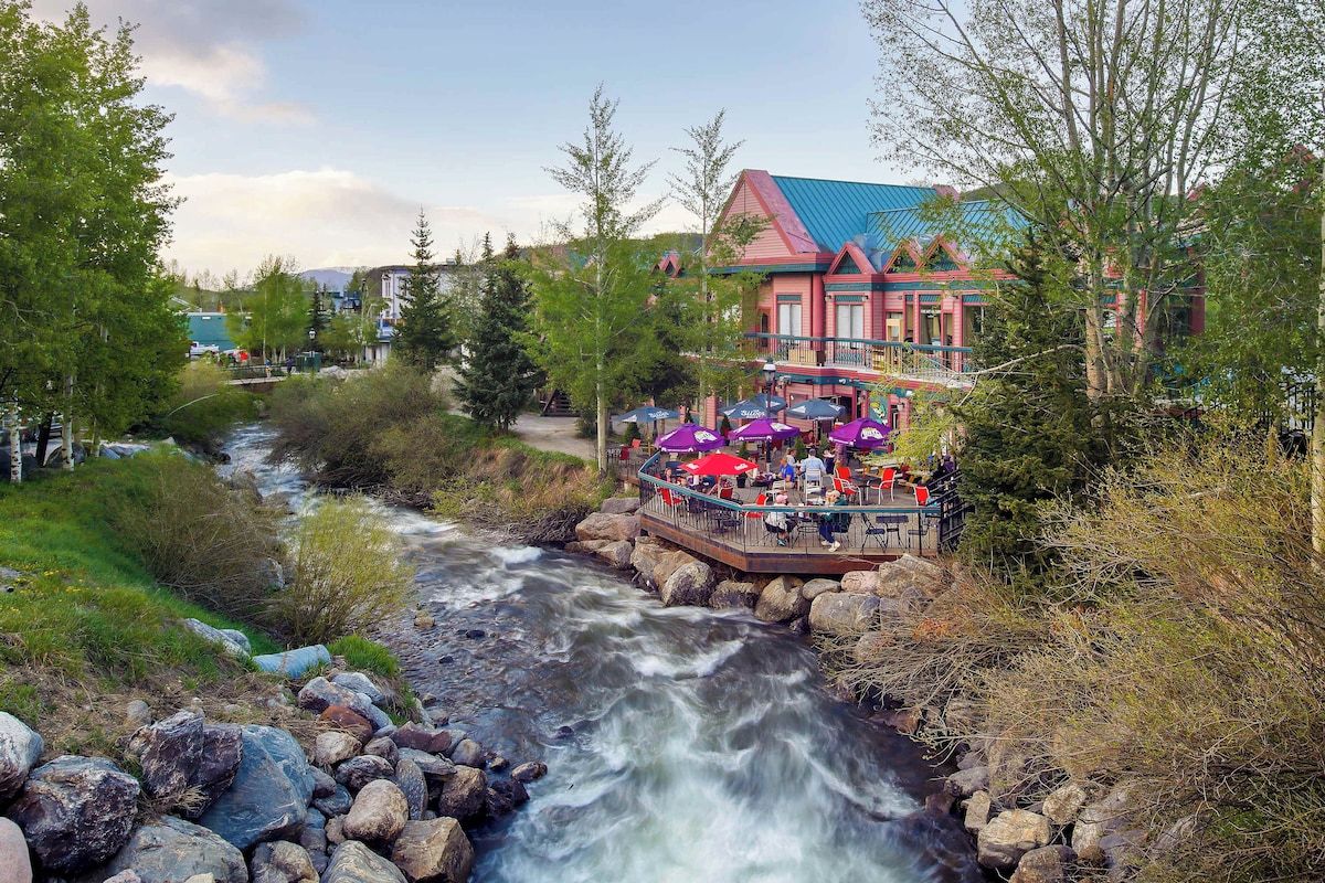Un restaurante junto al río con terraza al aire libre que ofrece vistas a un río serpenteante, rodeado de árboles y edificios.