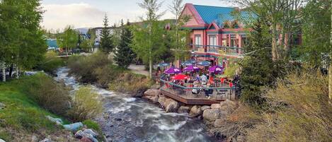 A riverside restaurant with outdoor seating overlooking a flowing river, surrounded by trees and buildings.