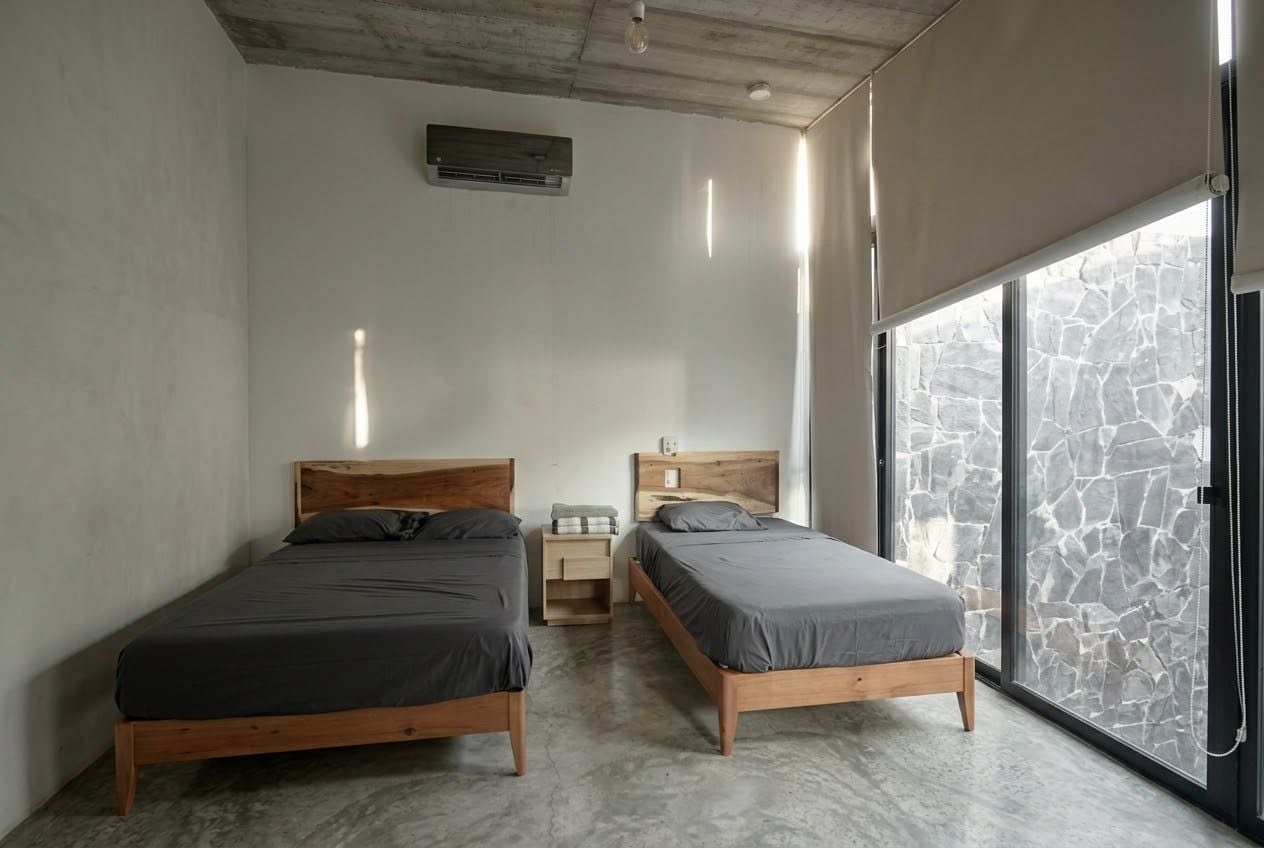 A bedroom with two wooden beds, a small bedside table, a wall-mounted air conditioner, and a large window with a stone wall view.