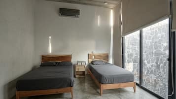 A bedroom with two wooden beds, a small bedside table, a wall-mounted air conditioner, and a large window with a stone wall view.