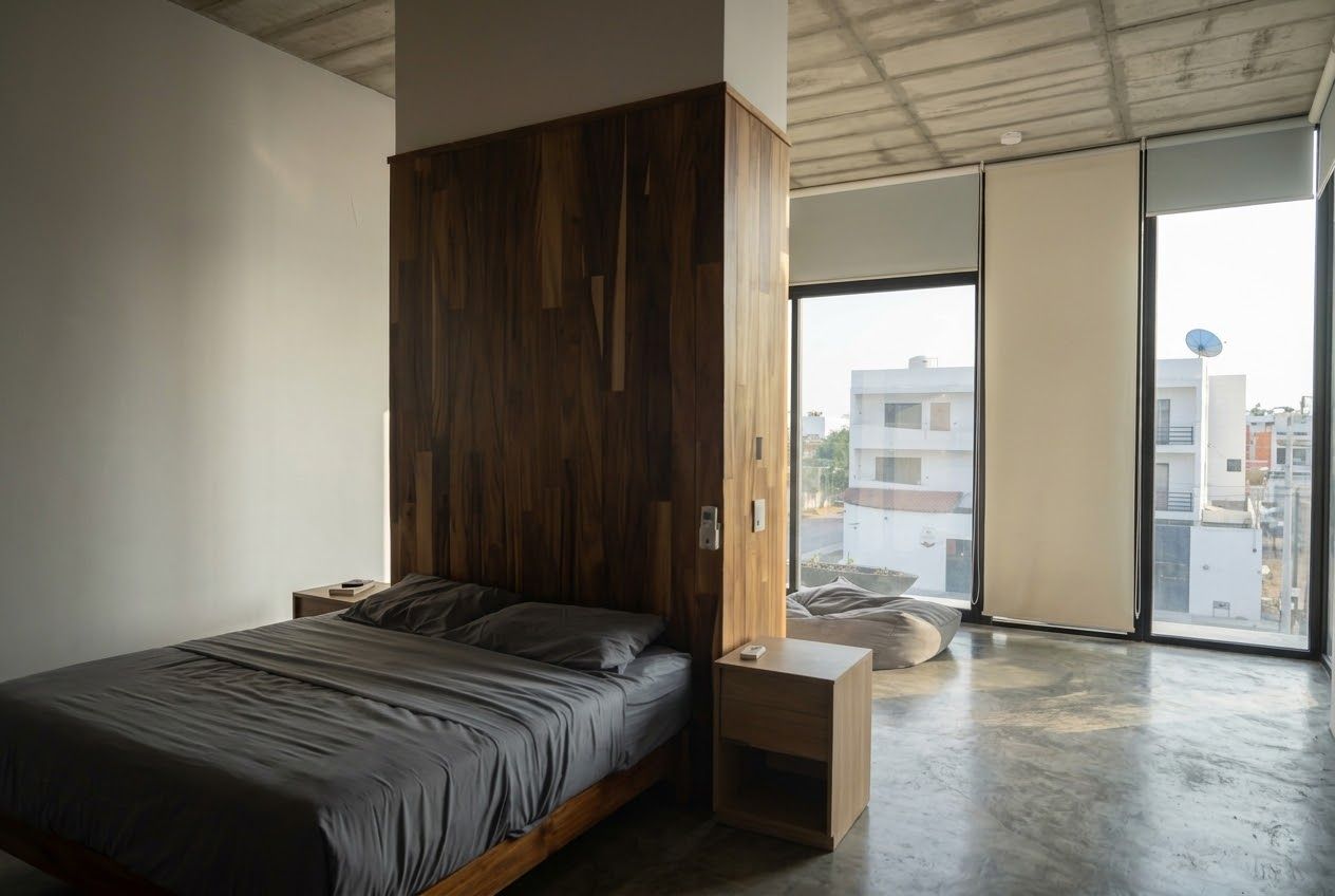 A modern bedroom with a wooden headboard, a bed with grey bedding, a bedside table, and a large window offering a city view.