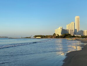 Uma praia com céu claro, mar calmo e uma fileira de prédios altos ao fundo.
