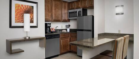 Cocina compacta con armarios de madera, refrigerador de acero inoxidable, microondas integrado y un pequeño comedor con banco.