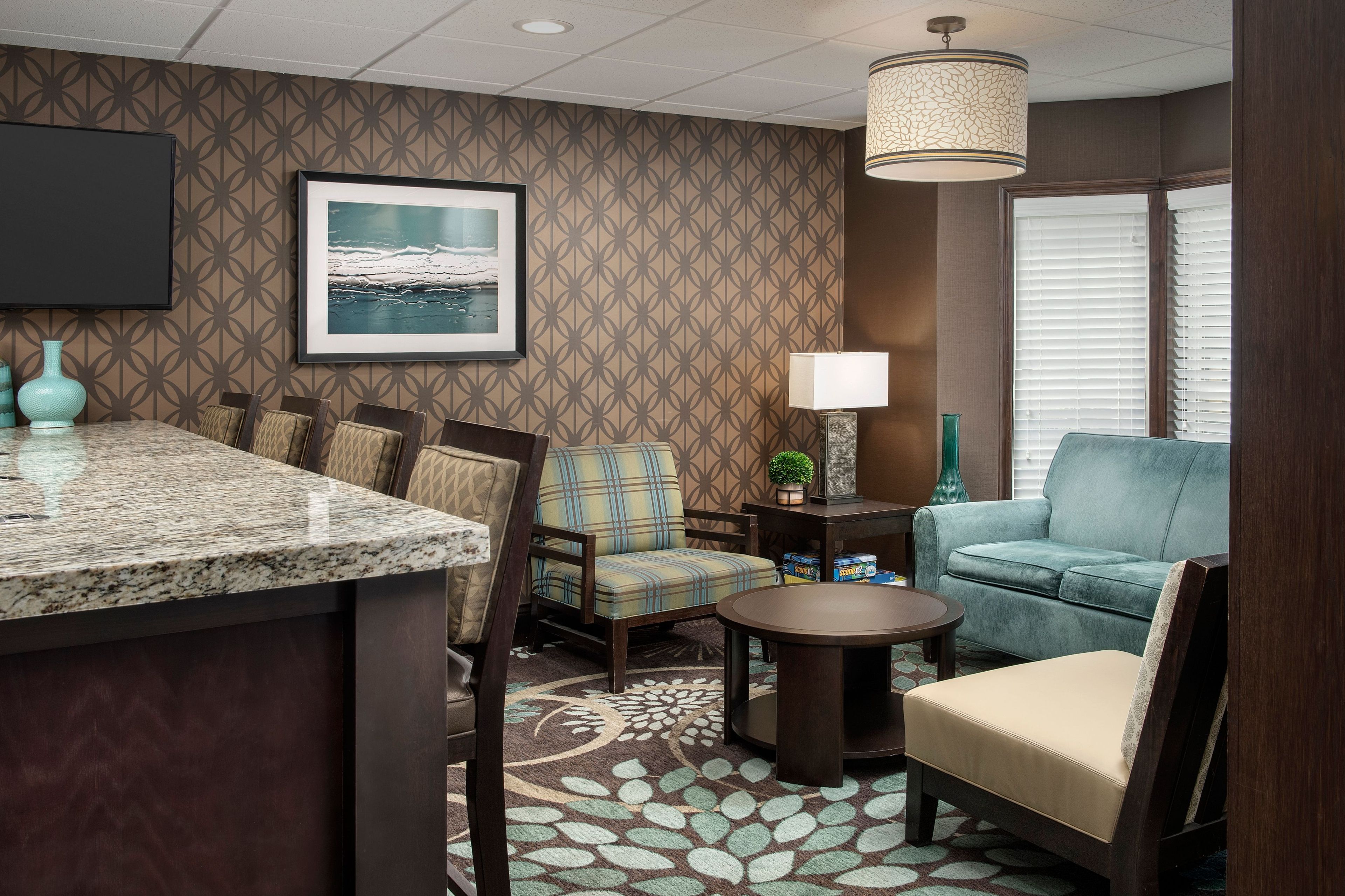 A hotel room with a kitchen area, a seating area with a sofa and chairs, a small table, a lamp, and a framed picture on the wall.