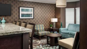A hotel room with a kitchen area, a seating area with a sofa and chairs, a small table, a lamp, and a framed picture on the wall.