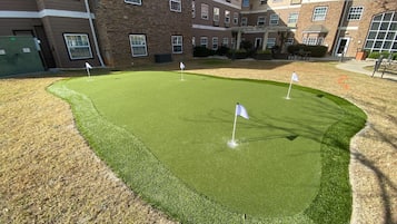 Un campo de golf artificial bien cuidado con banderas blancas que marcan los hoyos, rodeado por un edificio de ladrillo con numerosas ventanas.