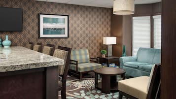 A hotel room with a kitchen area, a seating area with a sofa and chairs, a small table, a lamp, and a framed picture on the wall.