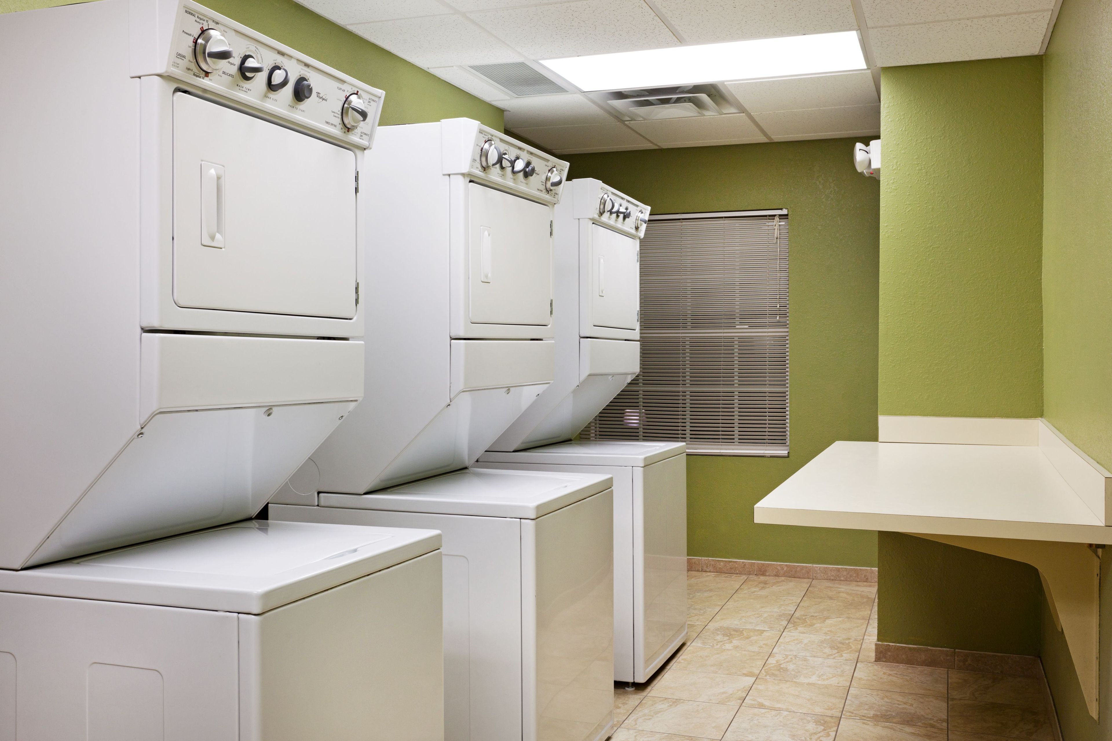 A laundry room with stacked washers and dryers, a window with blinds, and a small counter.