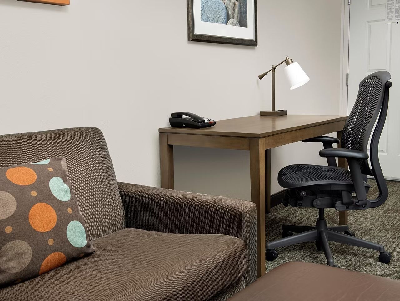 A room with a brown sofa, a black office chair, a wooden desk, a white desk lamp, and a telephone.