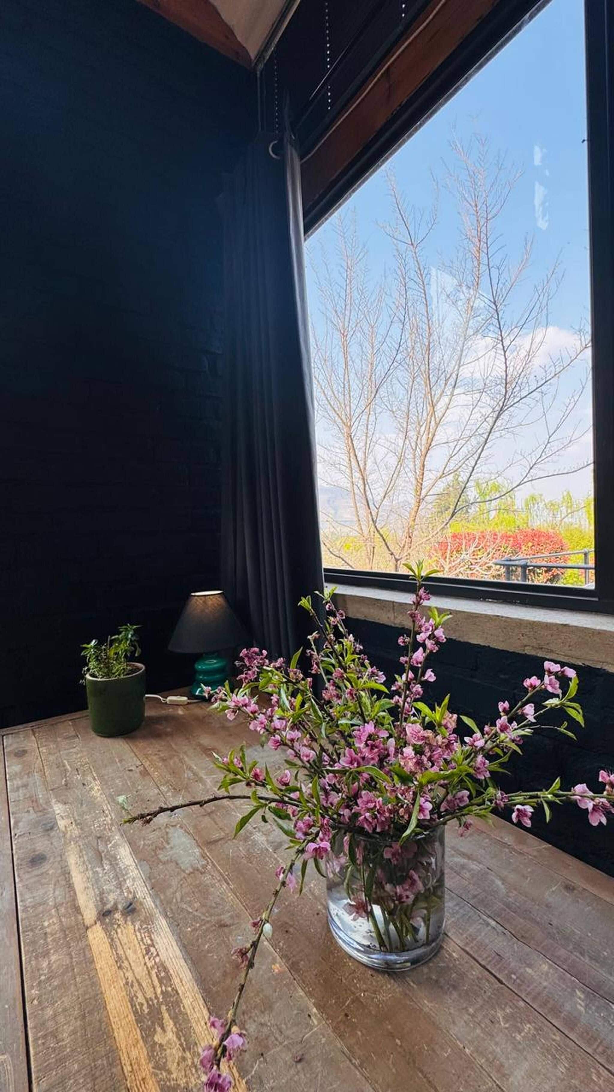 A room with a large window, a wooden table, a potted plant, and a vase with flowers.