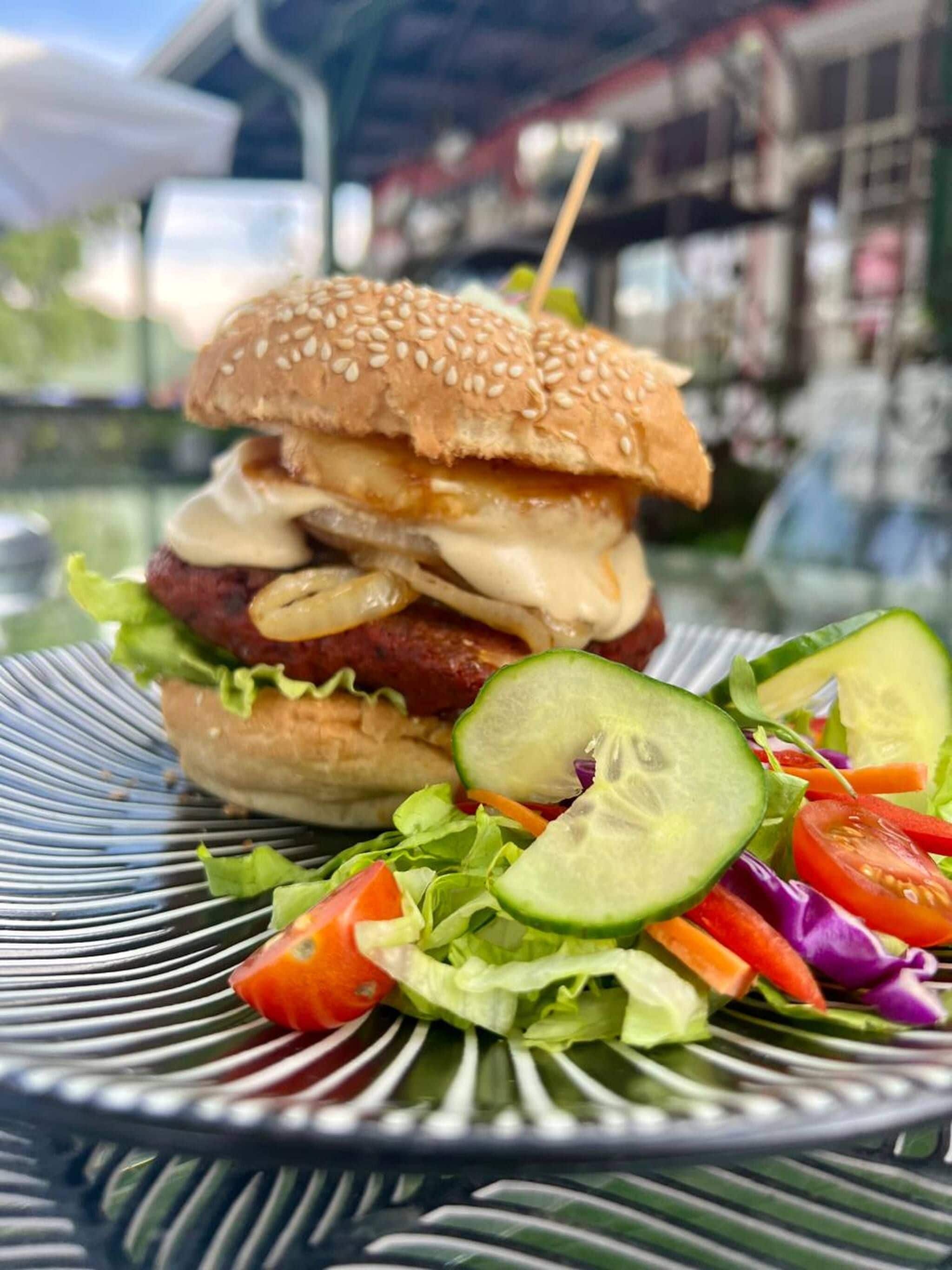 A burger with a sesame seed bun, cheese, and a side salad with lettuce, tomatoes, and cucumbers on a metal plate.
