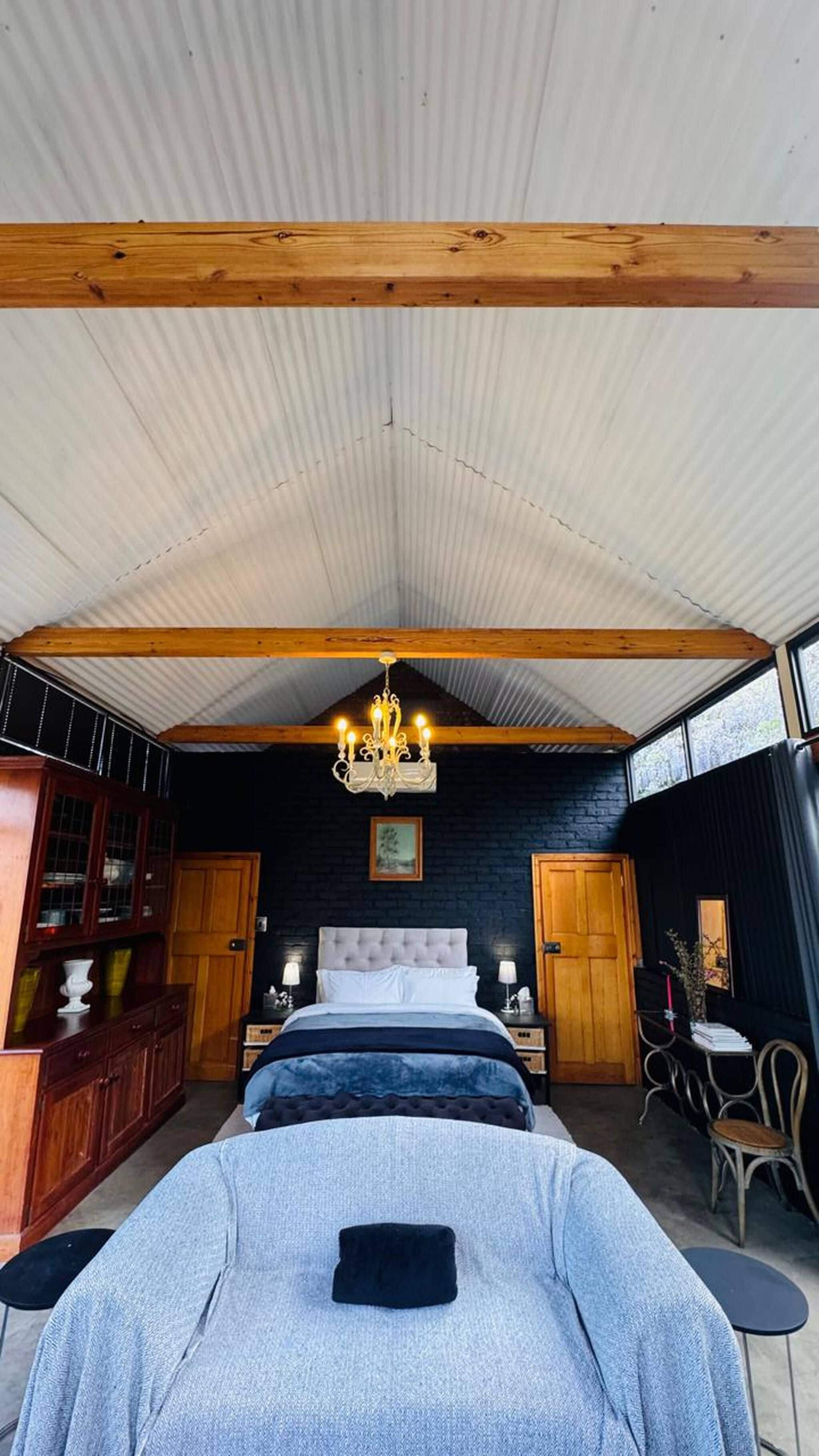 A bedroom with a large bed, a chandelier, wooden beams, a dark brick wall, and a window.