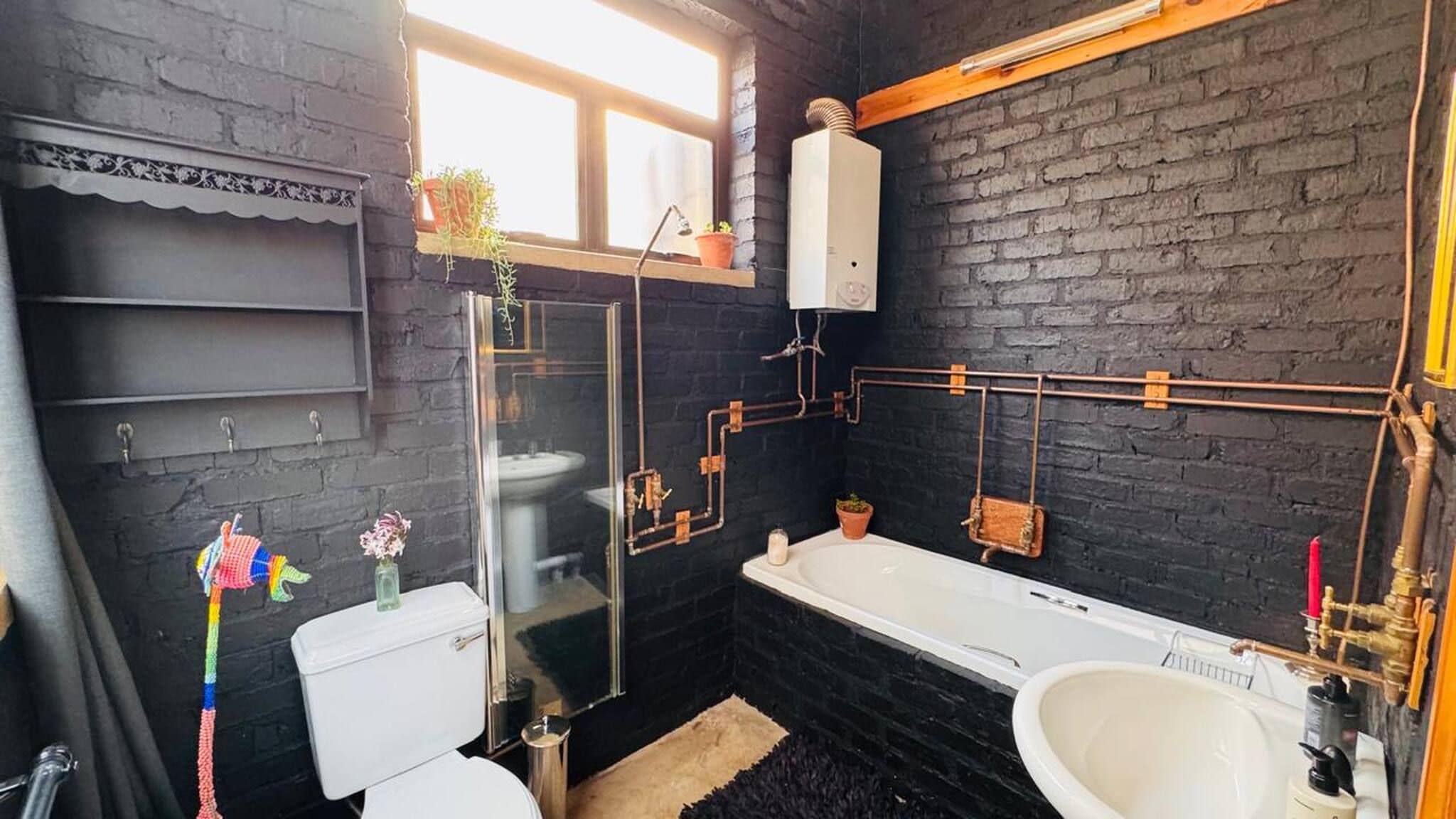 A bathroom with a black brick wall, a white toilet, a bathtub, and visible plumbing.