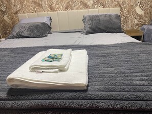 Una cama bien hecha con una toalla blanca doblada y un edredón blanco, un cabecero, dos almohadas y una colcha gris con textura.